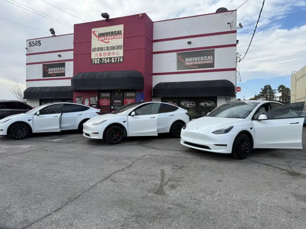 Three white Tesla cars are parked in front of a pink and gray business building with a sign, set against a cloudy sky.