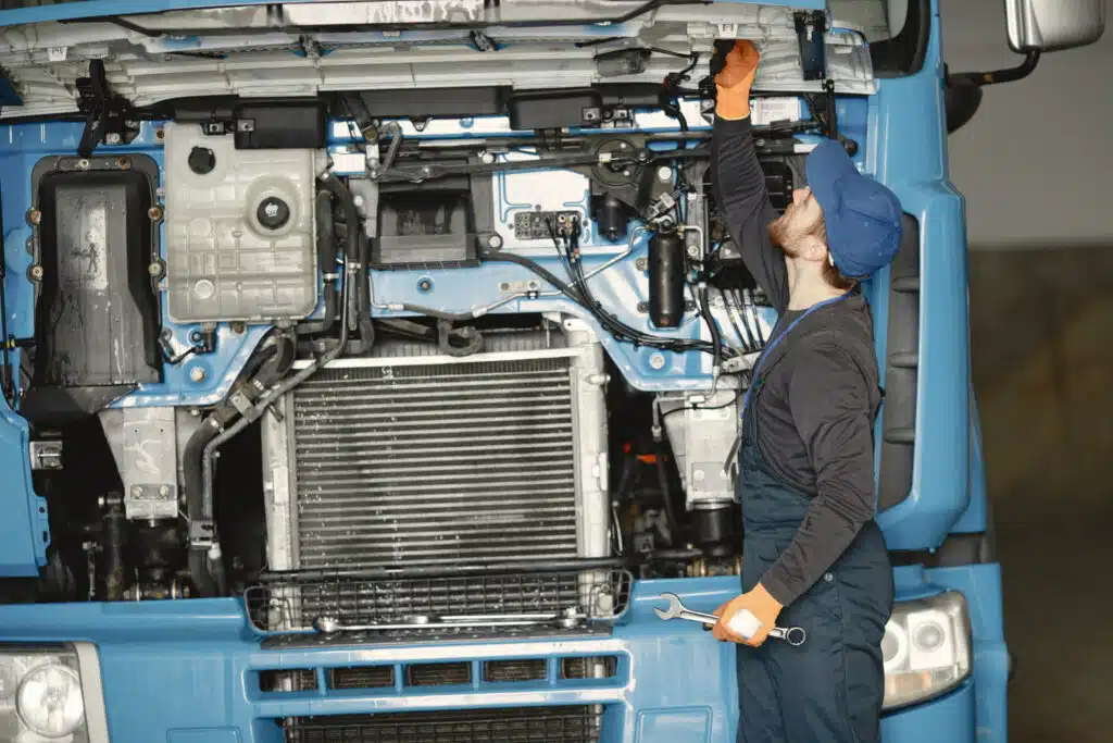 A mechanic in a blue cap and gloves inspects the engine of a large truck, holding a wrench while checking components under the hood.