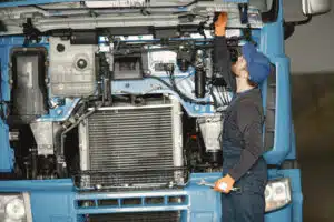 A mechanic in a blue cap and gloves inspects the engine of a large truck, holding a wrench while checking components under the hood.