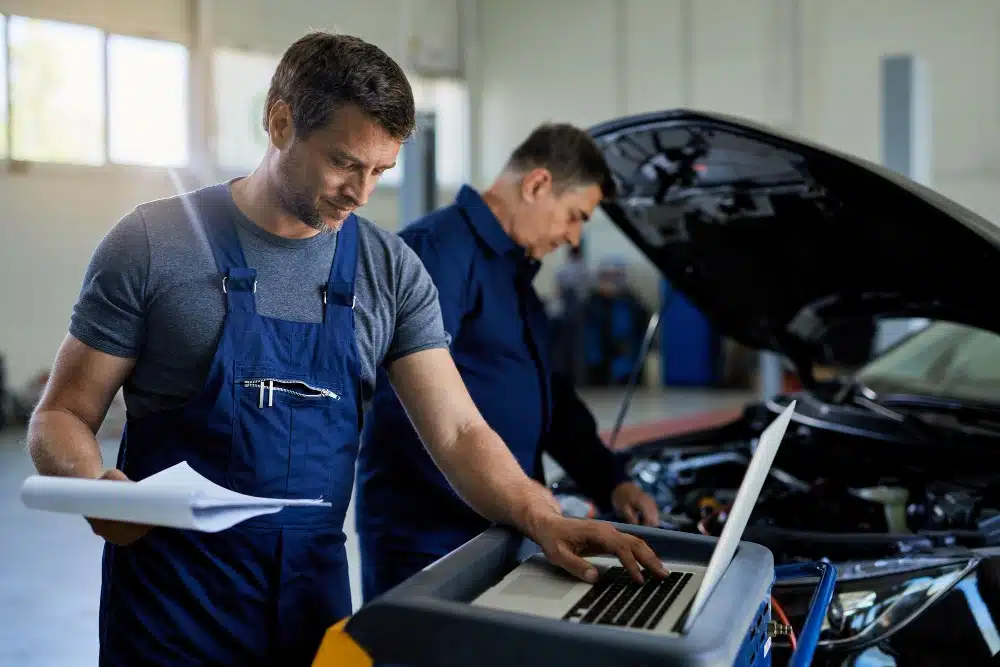 Two auto mechanics working in a garage, one using laptop by open hood at service