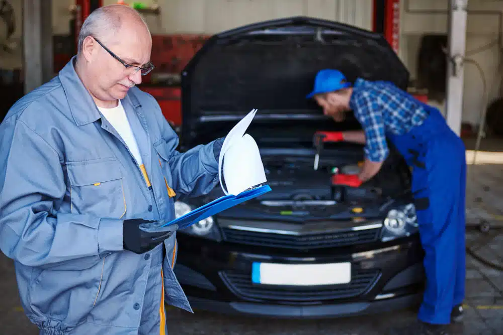 A mechanic reading a clipboard in a garage, while another mechanic works on the engine of a car.