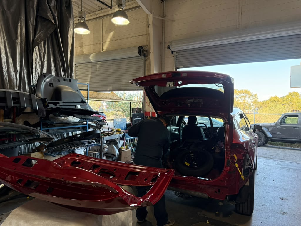 Technician working at the open rear of a red Kia SUV inside Universal Motorcars, with the rear bumper and body components removed.