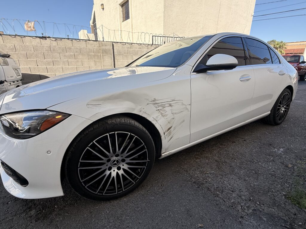 White Mercedes-Benz sedan with scraped and dented front-fender metal beside the wheel