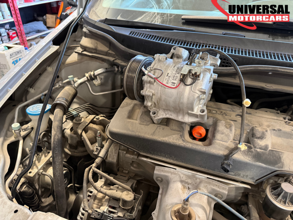 A/C compressor photographed above an open engine bay at Universal Motorcars.