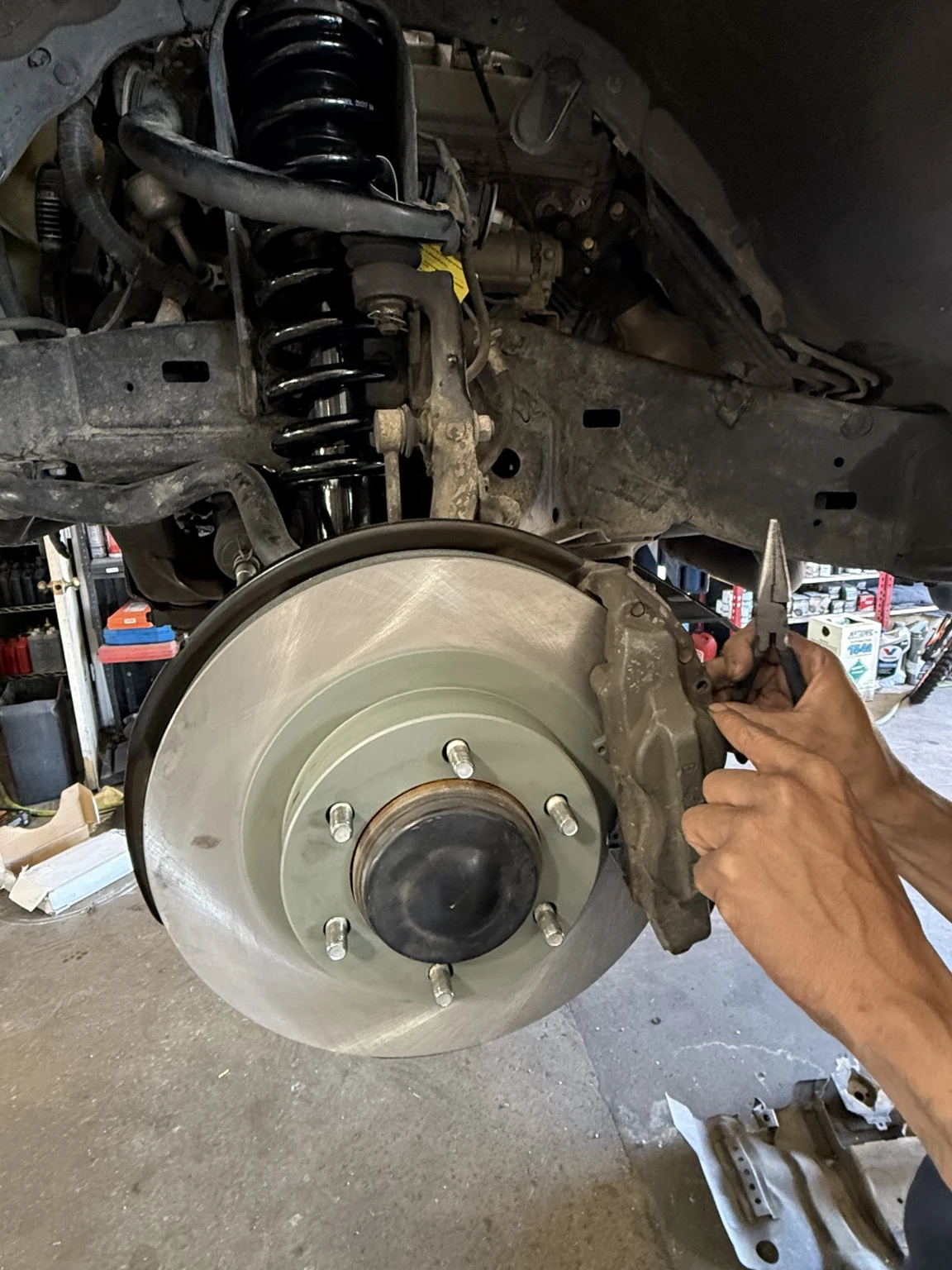 Hands work at an exposed brake caliper beside a brake rotor, coil spring and control arm.