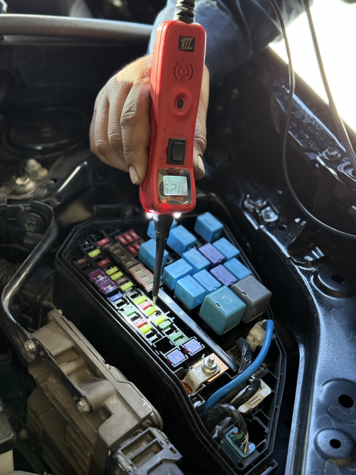 A hand holds an electrical test probe against a fuse in an under-hood fuse box.