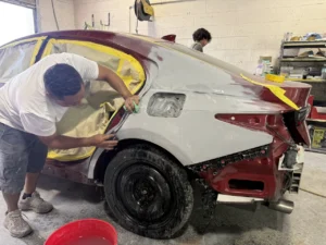 Technician sanding the rear quarter panel of a masked red sedan at Universal Motorcars.