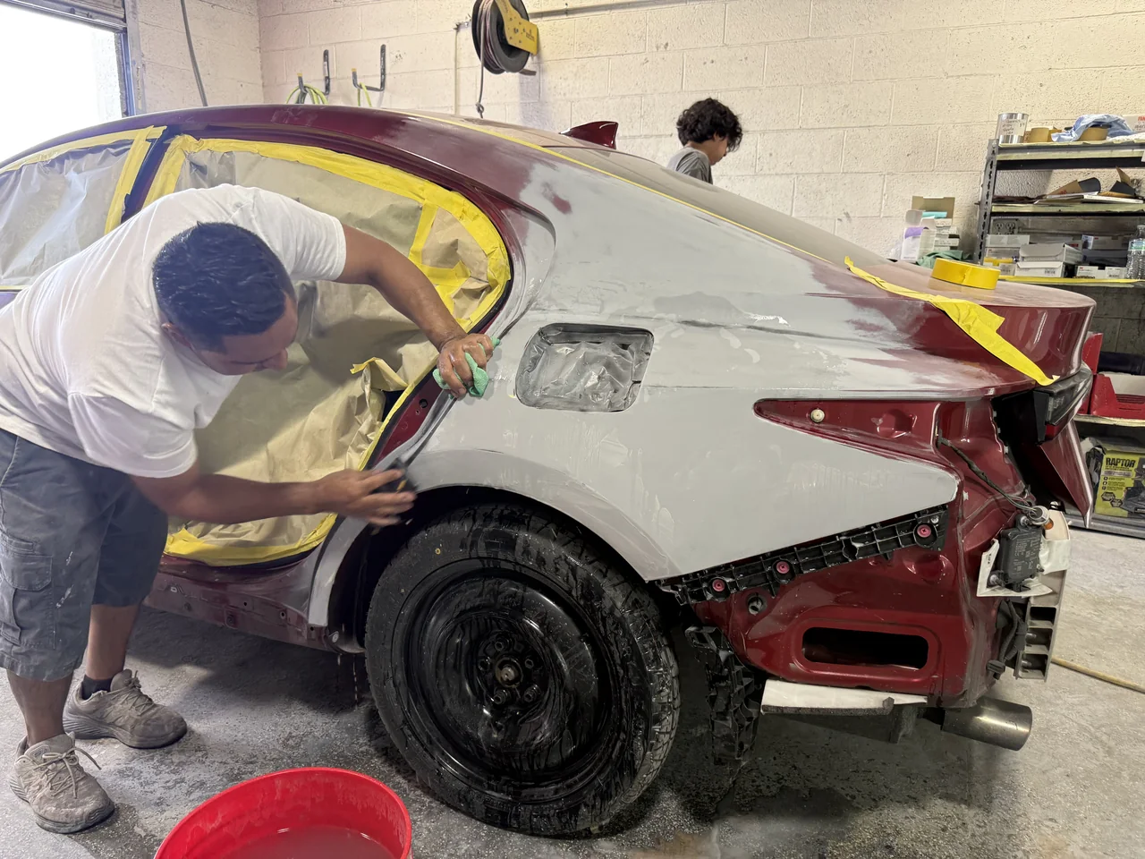 Technician sanding the rear quarter panel of a masked red sedan at Universal Motorcars.