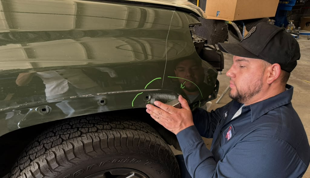Technician examining a marked vehicle body panel near the rear wheel in the workshop.