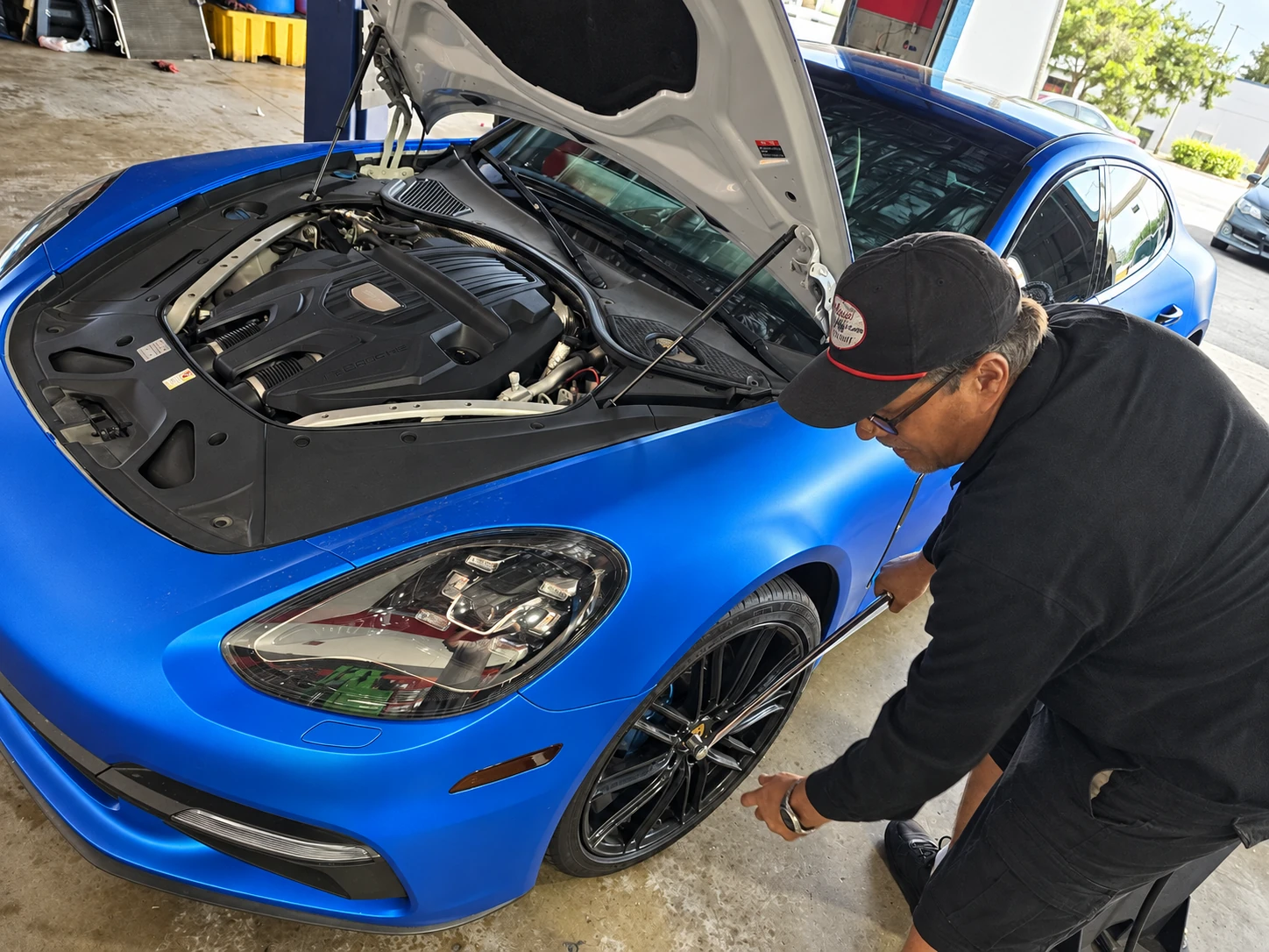 Technician beside the front wheel of a blue Porsche with the hood open at Universal Motorcars.