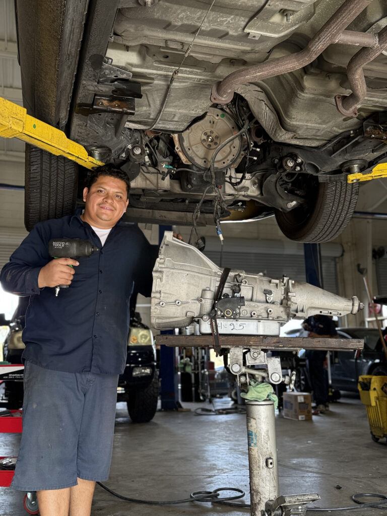 Universal Motorcars technician beside a removed transmission beneath a vehicle on a lift
