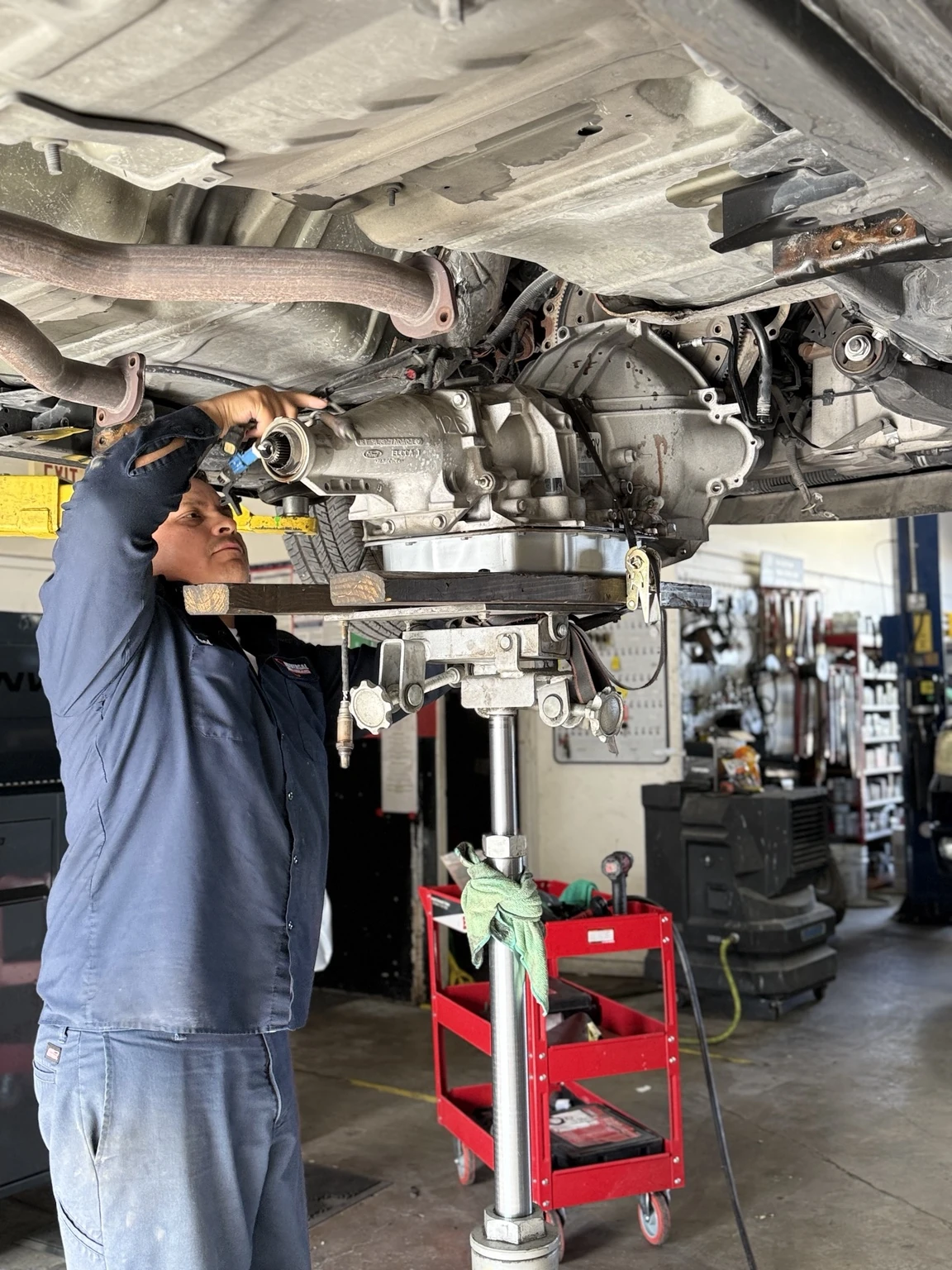 A technician works beneath a raised vehicle beside a transmission supported on a jack.