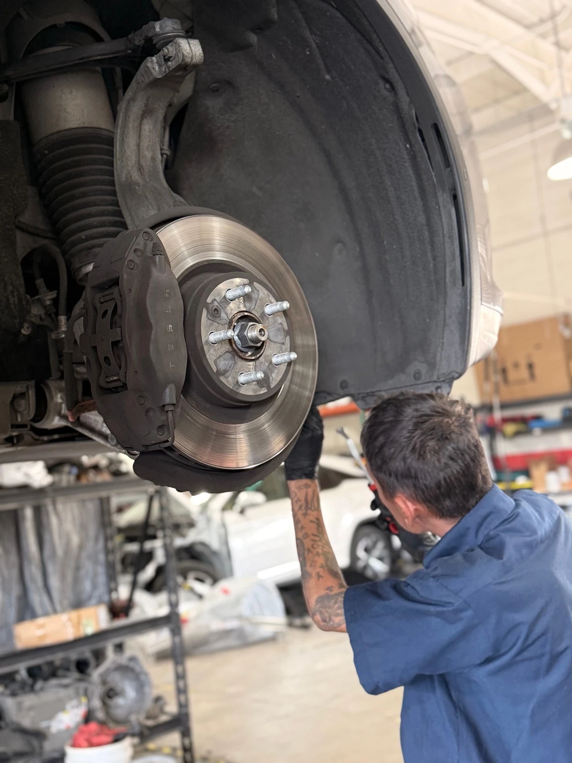 A technician works behind the exposed brake rotor and Tesla-branded caliper of a raised vehicle.
