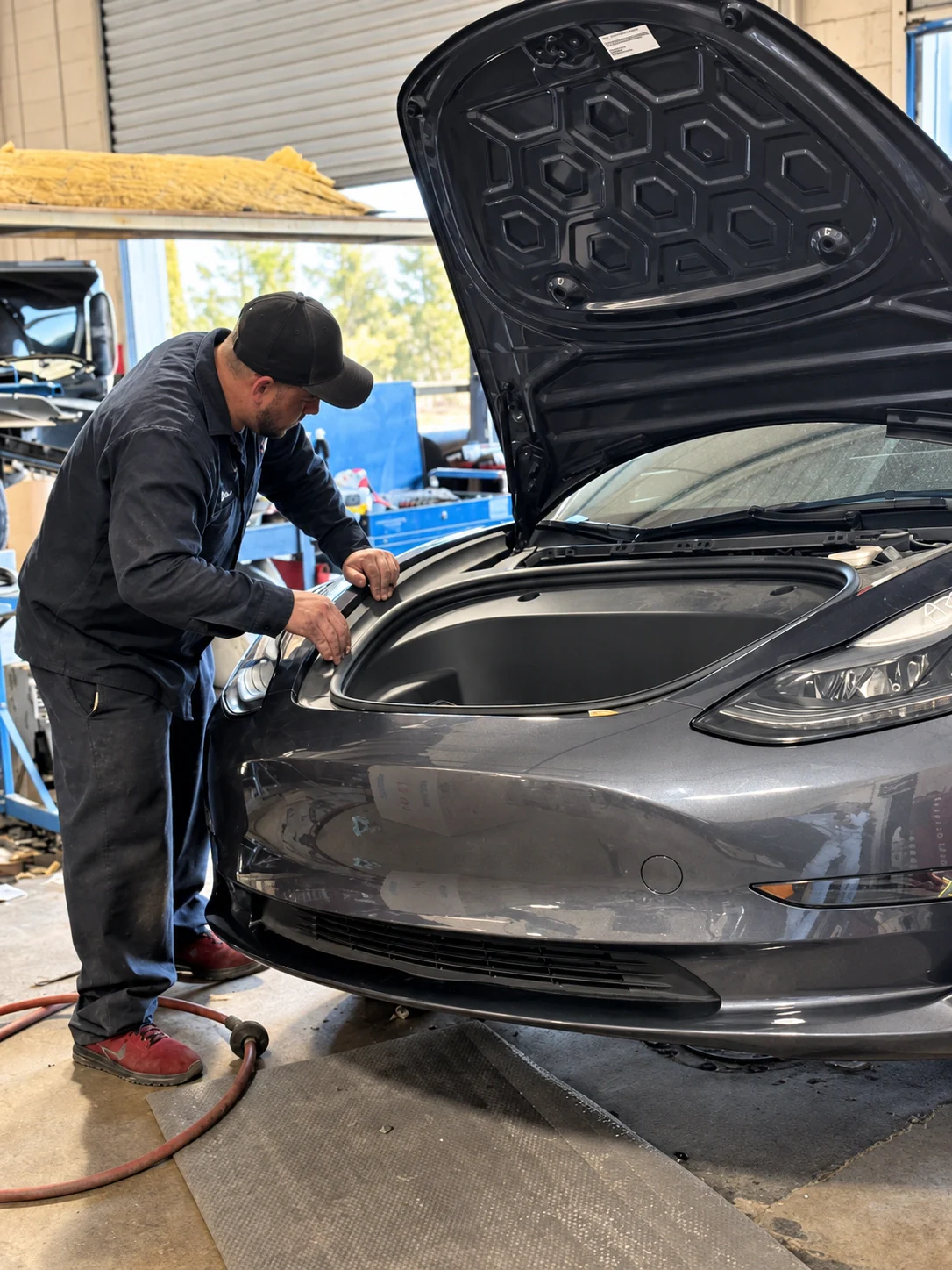 Technician working beside a gray Tesla with its front hood open in the workshop